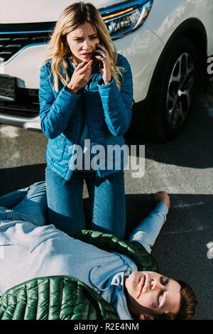 high angle view of scared young woman calling emergency while injured man lying on road after traffic accident Stock Photo