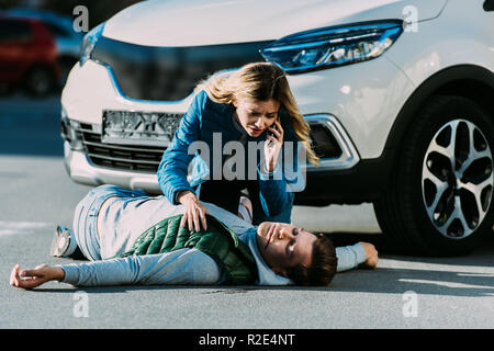 scared young woman calling emergency and looking at injured man on road after traffic accident Stock Photo