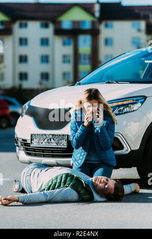 scared young woman calling emergency and looking at injured man lying on road after traffic collision Stock Photo