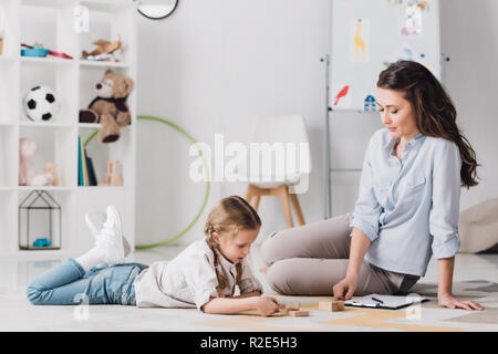 smiling psychologist sitting near child while she lying on floor and playing with wooden blocks Stock Photo