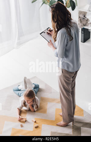 high angle view of psychologist with clipboard standing near child while she lying on floor and playing with wooden blocks Stock Photo