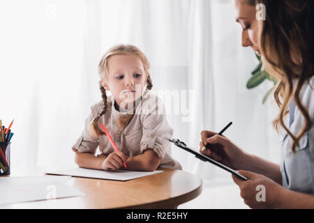 psychologist writing in clipboard while sitting near little drawing child Stock Photo