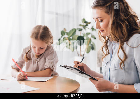 adult psychologist writing in clipboard while sitting near little drawing child Stock Photo