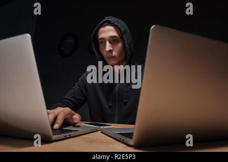 portrait of hacker in black hoodie using laptops at tabletop with smartphone, cyber security concept Stock Photo