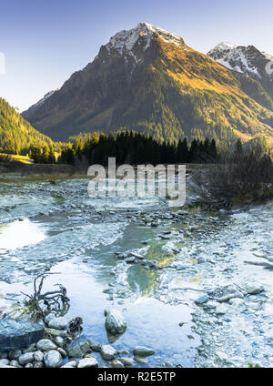 vertical rocky mountain landscape in early winter with snow-capped peaks and colorful forest and a mountain stream in the foreground with ice and rime Stock Photo