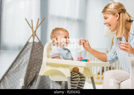 cheerful mom feeding son in highchair with baby food in nursery room Stock Photo