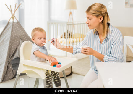 mother feeding son in highchair with baby food in nursery room Stock Photo