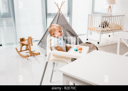 happy toddler sitting in baby chair in nursery room with toy cubes Stock Photo