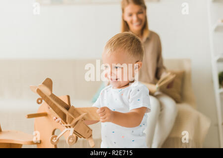 happy toddler playing with toy wooden toy plane in nursery room with mother on background