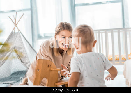 happy mother and toddler playing with toy wooden rocking horse chair in nursery room Stock Photo