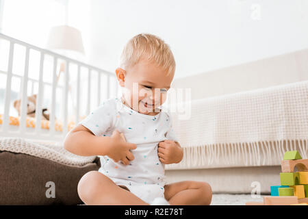 adorable toddler smiling and sitting on floor with colorful cubes in nursery room Stock Photo