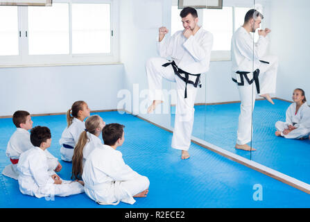 Happy young male coach explaining new maneuvers to children in karate class. Focus on the boy Stock Photo