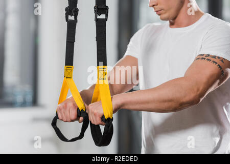 cropped image of tattooed athletic bodybuilder holding suspension straps in gym Stock Photo