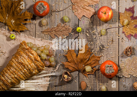 Homemade cornucopia filled up with white grapes. Dried maple leaves, persimmon fruit, apples, nuts, rye and wheat grains  on wooden surface. Stock Photo