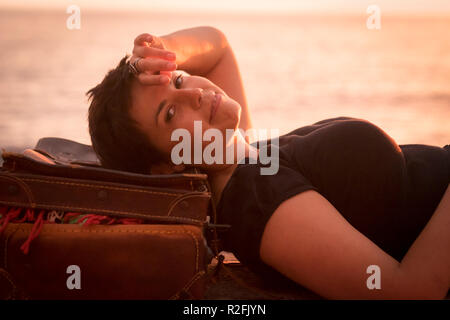 attractive and cheerful brunette lady curly black hair relaxed after the trip in outdoor on the coast. travel and destination concept for caucasian nice people. vintage luggage and modern lifestyle. ocean in background Stock Photo