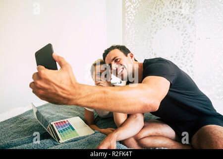 beautiful and cheerful caucasian young couple playing in relationship sitting on the white bed in bedroom and taking selfie with mobile phone. using laptop computer and internet working at home with joy and freedom Stock Photo
