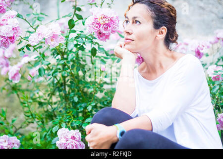 Beautiful wooman 40 years old look at her side thinking happy. Roses flower background. Nature outdoor colors. Stock Photo