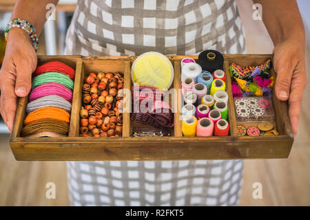 view from above woman hands taking a wooden box full of stuffs and articles for a tailor to make and repair clothes and dresses. colorful background with beads, and textile related fabric. people at work Stock Photo