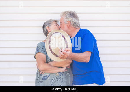 Valentine's day concept with elderly beautiful senior couple kissing hidding with a beige hat with a white wood background wall behind them. love and forever life together with caucasian couple man and woman happy Stock Photo