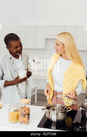 mature blonde woman cooking dinner while african american man drinking coffee Stock Photo