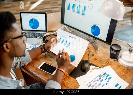 Young african man working with analitycs sitting at the beautiful working place with computer, laptop and documents. View from above Stock Photo