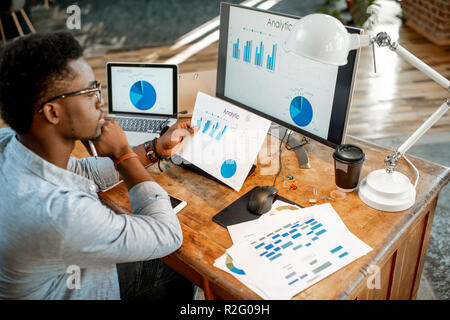 Young african man working with analitycs sitting at the beautiful working place with computer, laptop and documents. View from above Stock Photo