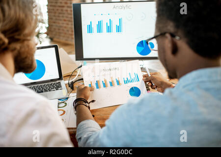 Two male coworkers working with analitycs sitting together at the working place with computer, laptop and documents. View from behind focused on the documents Stock Photo
