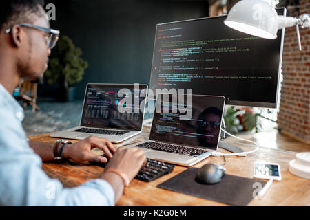 Young african male programmer writing program code sitting at the workplace with three monitors in the office. Image focused on the screen Stock Photo