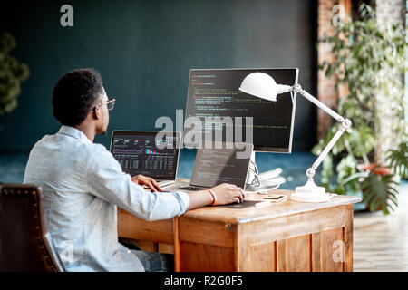 Young african male programmer writing program code sitting at the workplace with three monitors in the office. Image focused on the screen Stock Photo