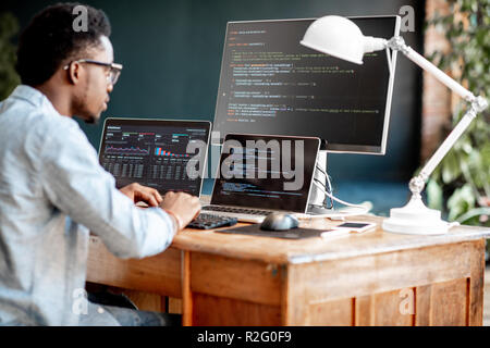 Young african male programmer writing program code sitting at the workplace with three monitors in the office. Image focused on the screen Stock Photo