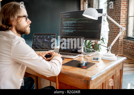 Young male programmer writing a program code sitting at the workplace with three monitors in the office. Image focused on the screen Stock Photo