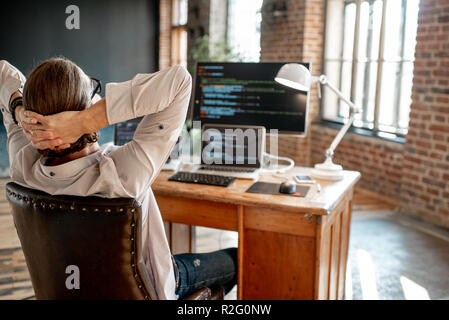 Young male programmer relaxing during the work with program code sitting at the workplace with three monitors in the office Stock Photo