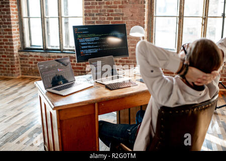 Young male programmer relaxing during the work with program code sitting at the workplace with three monitors in the office. Image focused on the screen Stock Photo