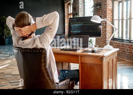 Young male programmer relaxing during the work with program code sitting at the workplace with three monitors in the office. Image focused on the screen Stock Photo