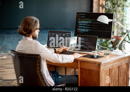 Young male programmer writing a program code sitting at the workplace with three monitors in the office Stock Photo