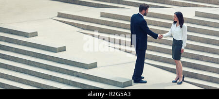 Panoramic web banner photograph of caucasian man businessman and an Asian woman businesswoman shaking hands on a deal standing on modern city steps Stock Photo