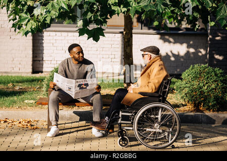 senior disabled man in wheelchair and african american man spending time together and reading newspaper on street Stock Photo