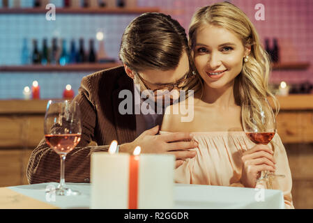 smiling girl with wine glass looking at camera while her boyfriend kissing her shoulder at table in restaurant Stock Photo