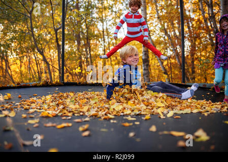 Three children playing on a trampoline covered in autumn leaves, United States Stock Photo