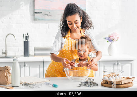 smiling african american mother helping daughter breaking egg for preparing dough in kitchen