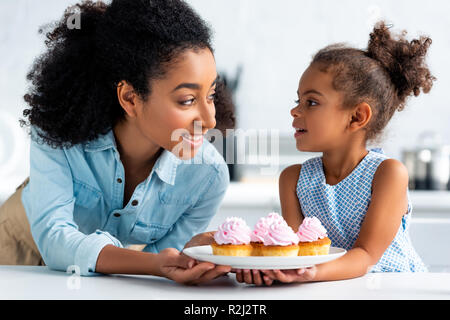 smiling african american mother and daughter holding homemade cupcakes and looking at each other in kitchen Stock Photo