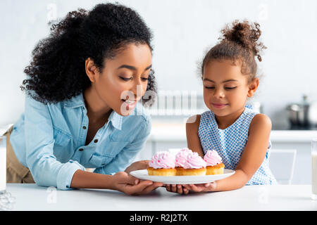 african american mother and daughter looking at homemade cupcakes in kitchen Stock Photo