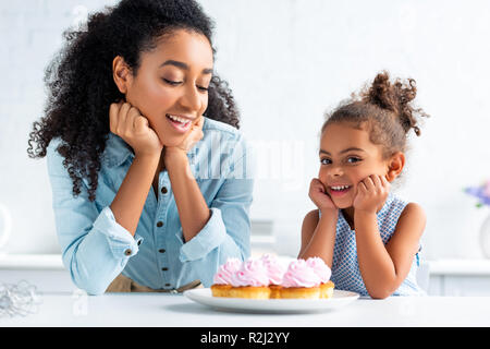 happy african american mother and daughter resting chins on hands near cupcakes on table in kitchen Stock Photo