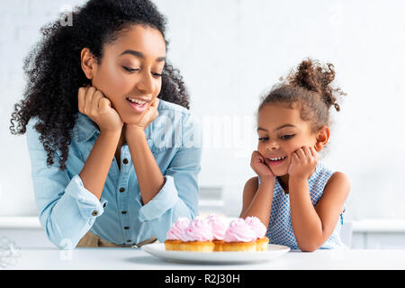 cheerful african american mother and daughter resting chins on hands and looking at cupcakes on table in kitchen Stock Photo