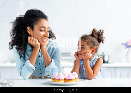 cheerful african american mother and daughter resting chins on hands and looking at each other, cupcakes on table in kitchen Stock Photo