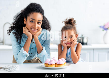 cheerful african american mother and daughter resting chins on hands and looking at camera, cupcakes on table in kitchen Stock Photo