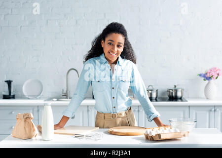 smiling attractive african american girl leaning on kitchen counter and looking at camera Stock Photo