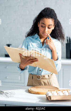 pensive beautiful african american girl reading cookbook in kitchen Stock Photo