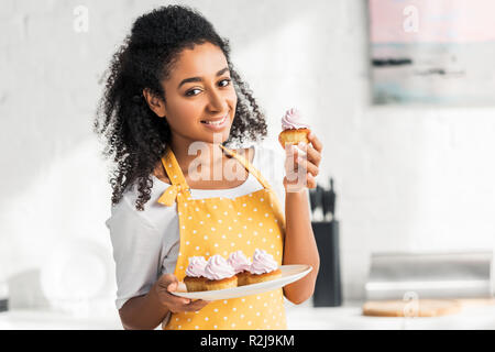 smiling attractive african american girl in apron holding homemade cupcakes and looking at camera in kitchen Stock Photo