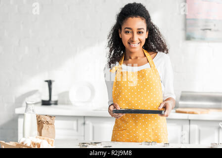 smiling attractive african american girl in apron holding tray with unbaked cookies in kitchen Stock Photo